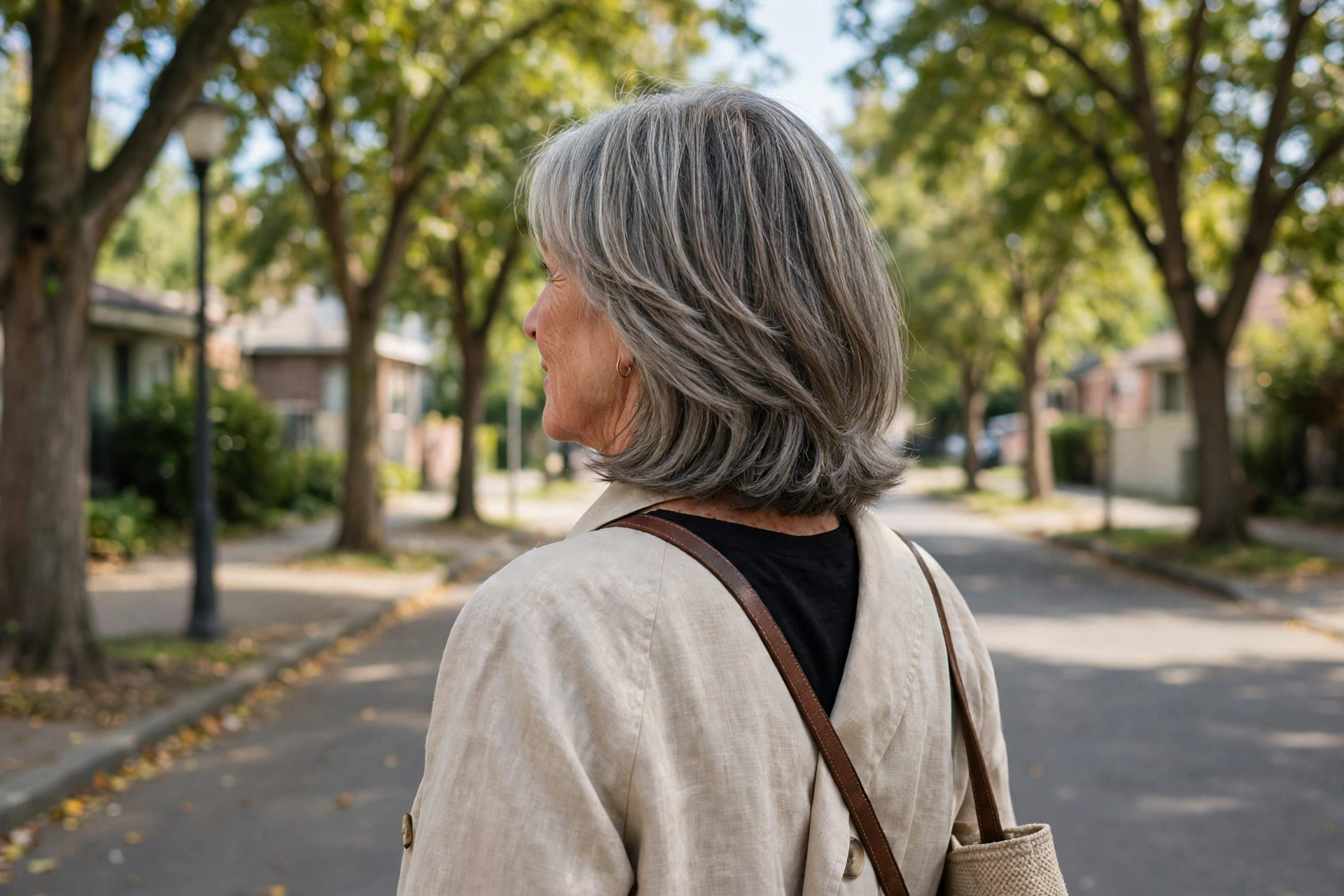 late-sixties woman, graceful collarbone-length cut with soft blended layers, bright tree-lined street, from behind at three-quarter angle