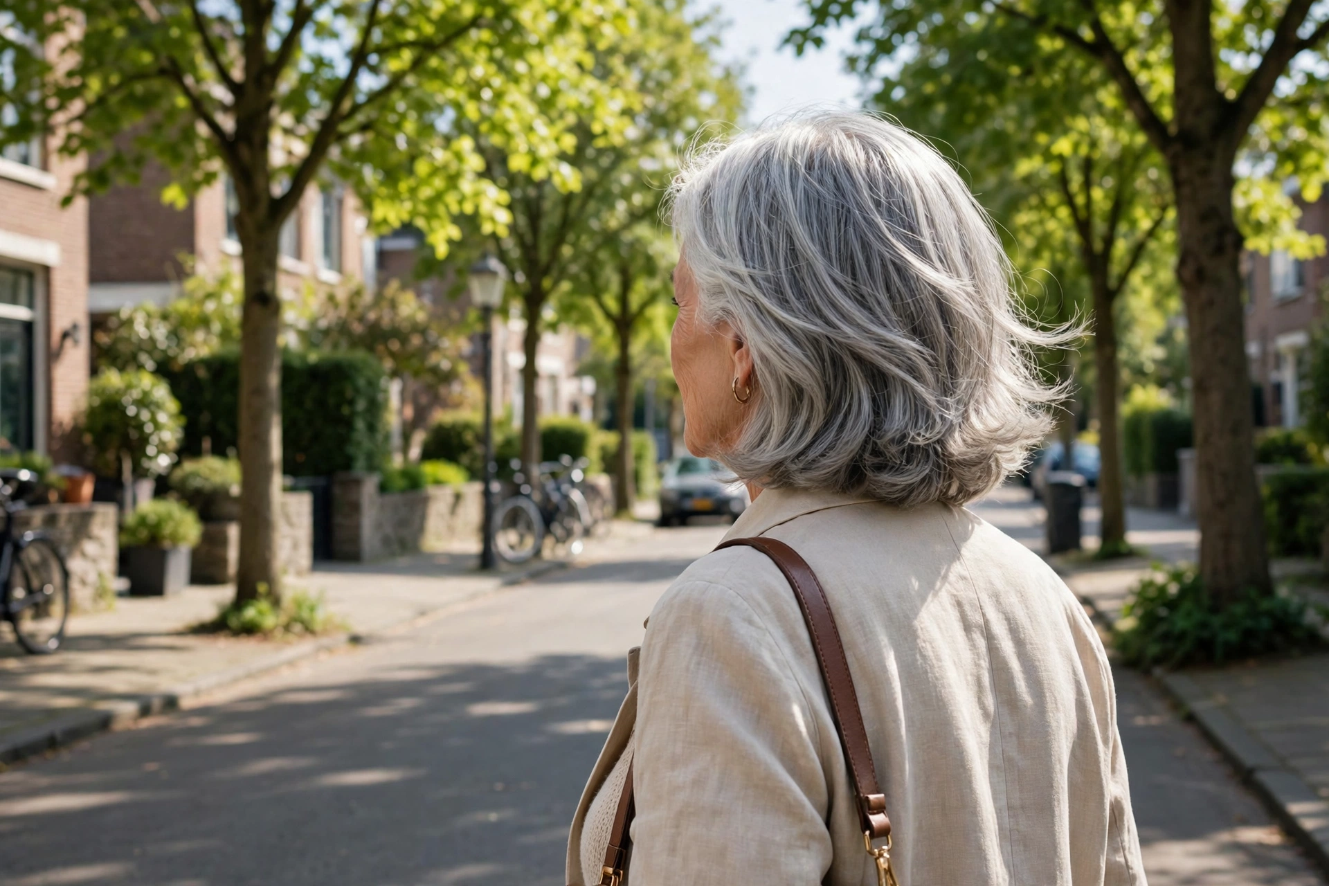 A woman in her early seventies with a collarbone cut and gentle movement, walking along a bright quiet street lined with trees in daylight, from behind at a three-quarter angle, relaxed elegant mood.