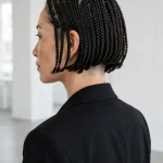 A woman with a chin-length braided bob and a closely cut undercut visible at the nape, standing in a bright modern studio with white walls, wearing a black blazer, photographed from a three-quarter back angle, clean bright style.