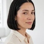 A woman with a glossy, chin-length braided bob with slicked roots and a blunt even edge, standing in a bright modern studio with white walls, wearing a cream blouse, photographed from a three-quarter front angle, clean bright style.