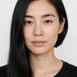 A woman with an asymmetrical bob, one side clearly longer than the other, sleek dark hair, photographed front-facing in a bright studio with a plain background, wearing a black top.