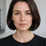 A woman with a crisp chin-length bob with tucked-under ends, smooth dark brown hair, wearing a black top and gold hoop earrings, photographed front-facing in a bright studio.