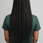 A woman with waist-length box braids flowing down her back, photographed from behind in a bright studio with a neutral background, showing the dramatic full length of the plaits.