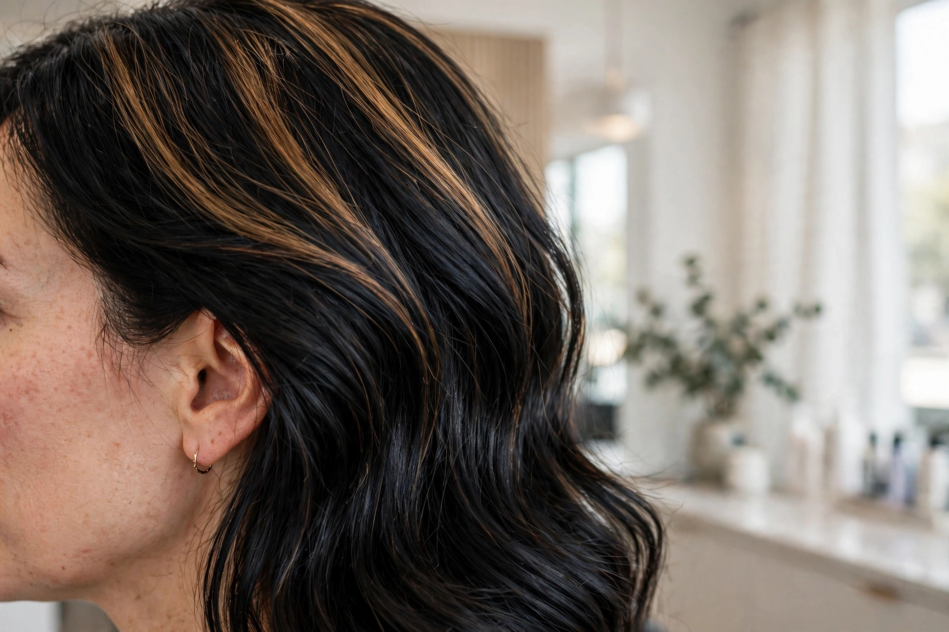 Close-up side view of a woman's black hair showing honey lift highlights through the upper sections, in a bright modern salon setting, soft natural light, clean bright style.