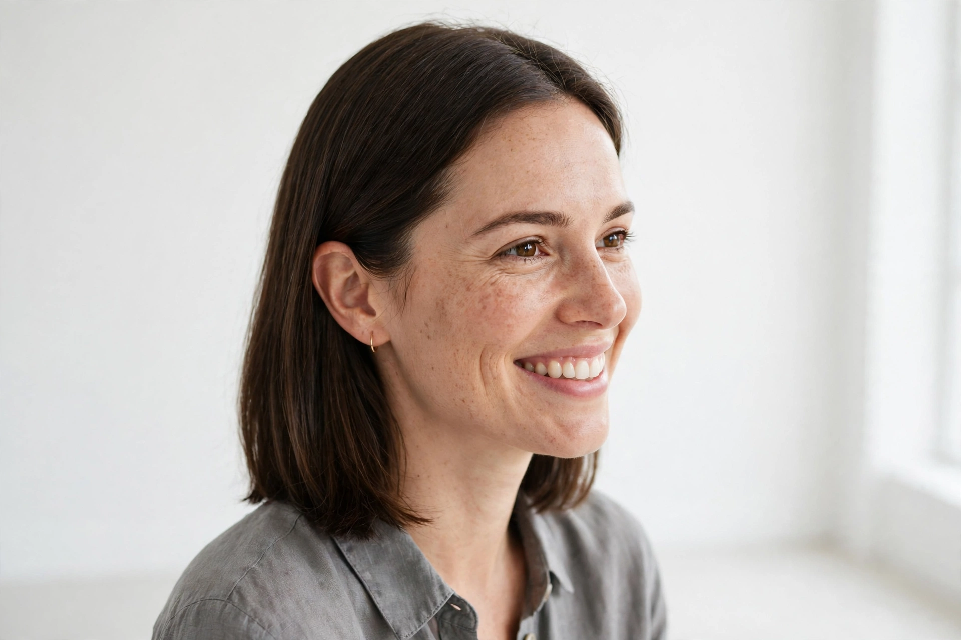 A woman with medium hair in a shoulder cut with a tuck-and-go length smiling in a bright minimalist studio, hair tucked neatly behind one ear, wearing a gray blouse, even daylight, three-quarter angle, clean bright style.