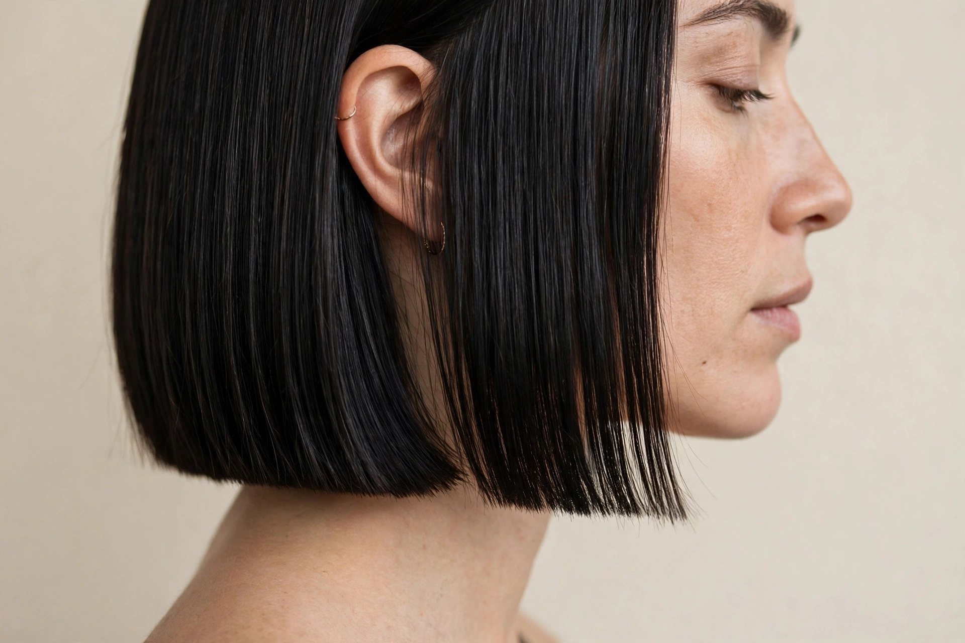 Close-up side profile of a woman with a blunt cut in sleek black hair, photographed in soft studio light against a pale beige background, with the razor-sharp, straight-across hemline in focus.