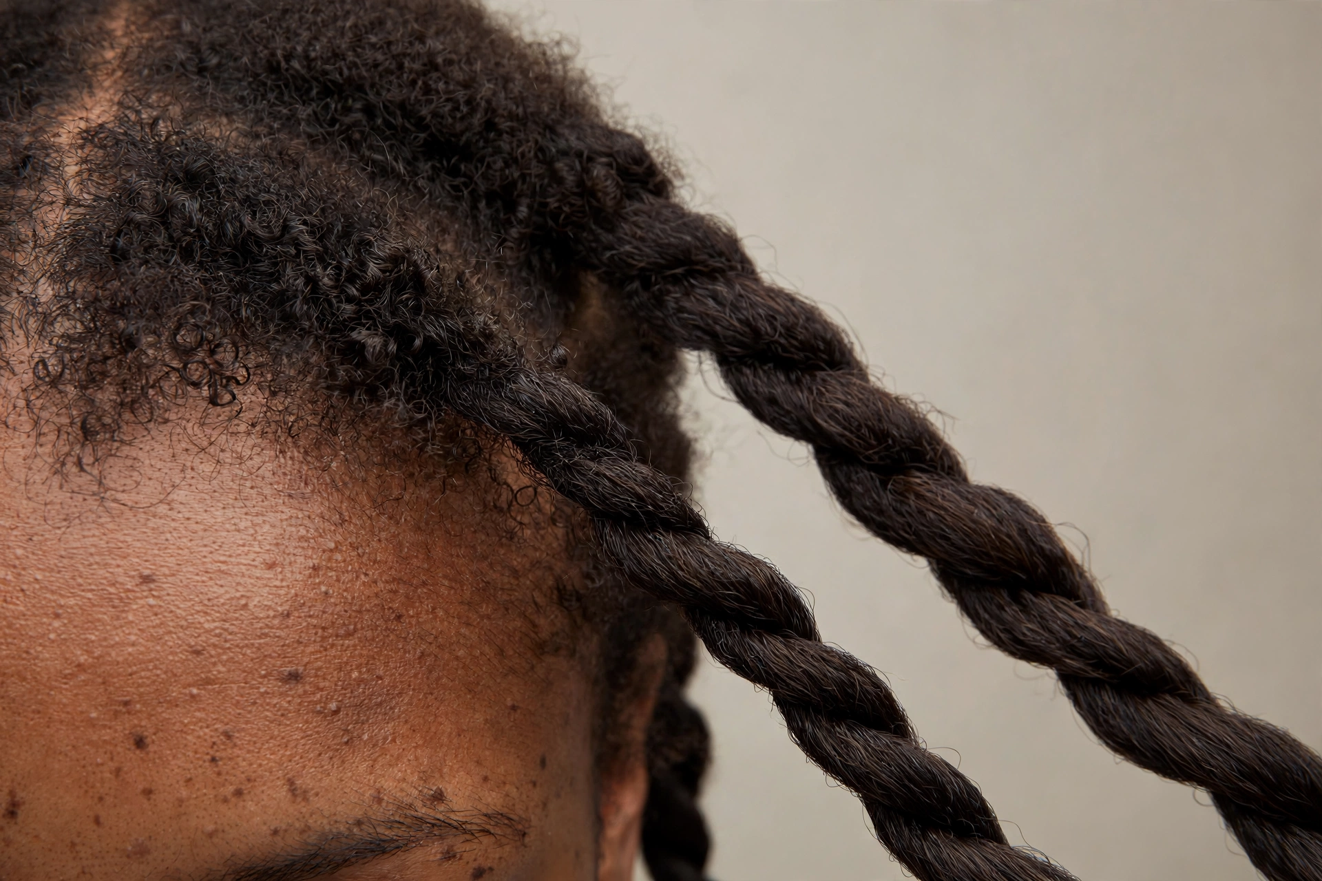 Close-up of two-strand twists on transitioning hair with natural curly/coily roots visible at the scalp, photographed in soft studio light against a neutral background, partial face visible.