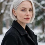 A woman with an icy platinum sleek bob and a flawless smooth finish, wearing a dark wool coat, standing outdoors in soft winter daylight with snow-dusted trees in soft focus behind her, three-quarter front angle, clean bright style.